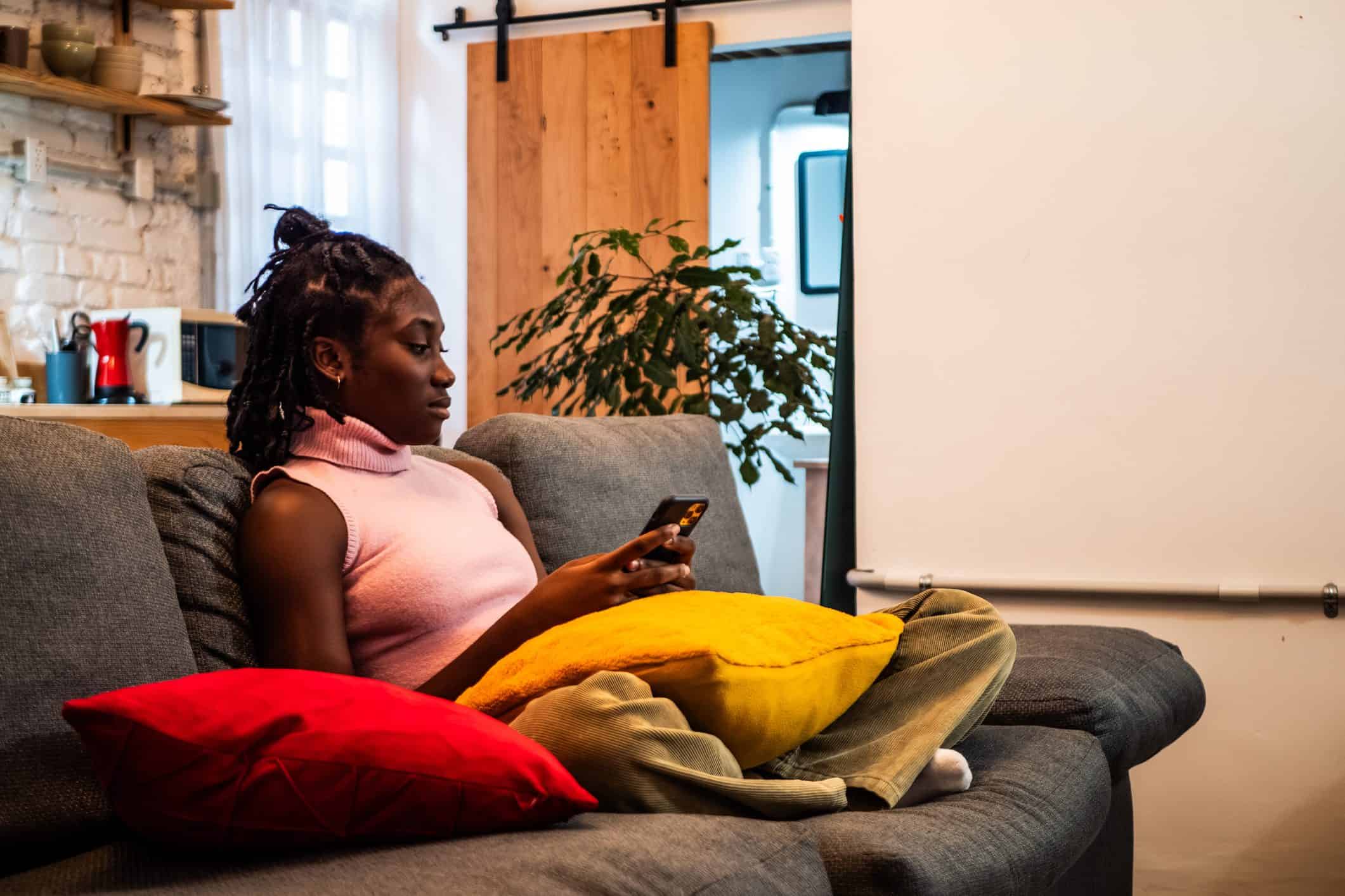 A young Black woman sits on her sofa, using her cell phone.