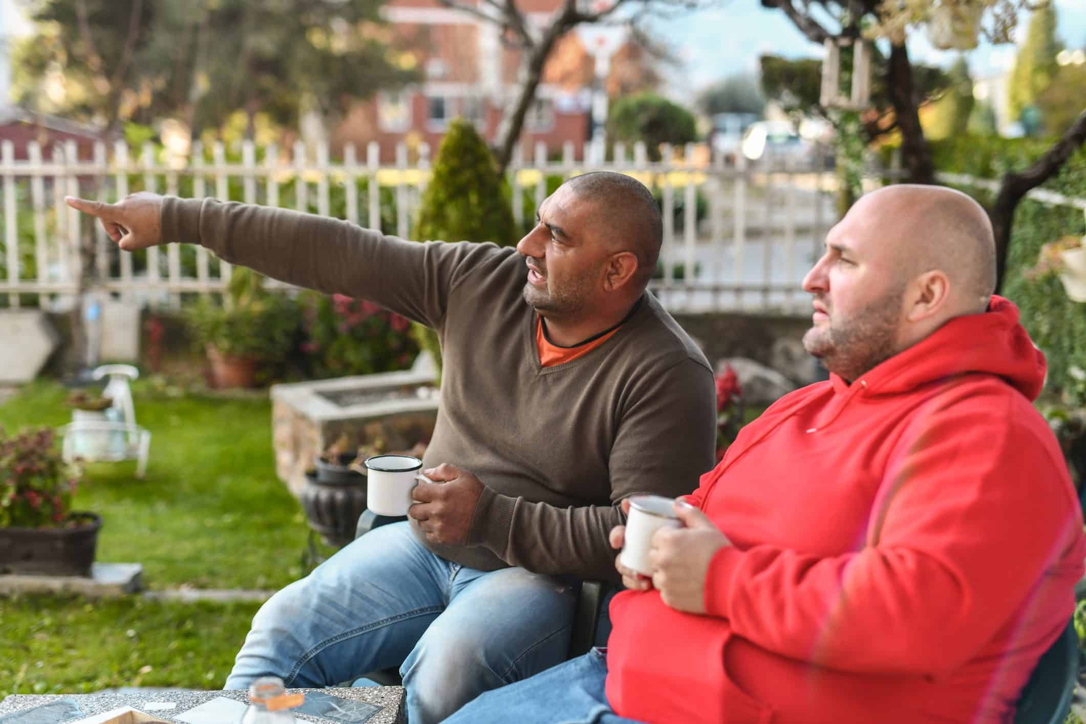 Two men sit on a front porch, drinking coffee.